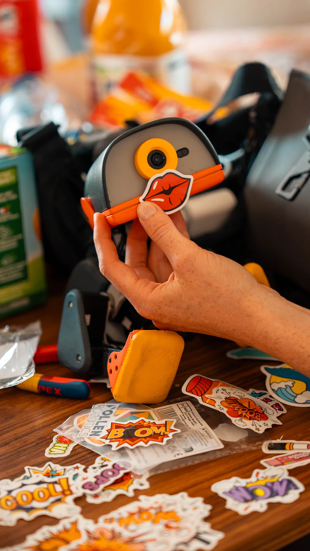 A hand sticking a lips sticker onto a MACRODUCK's beak, sticker sheets spread on the table